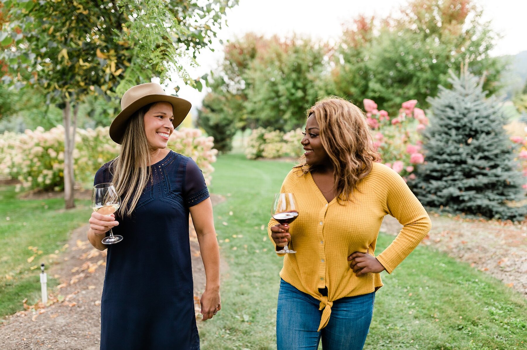 Two women, each holding a wine glass, walk through a vineyard.