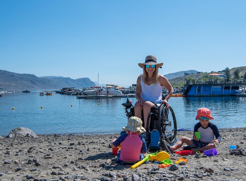 Woman in wheelchair and kids playing in the sand at Bruker Marina beach with boats in background.
