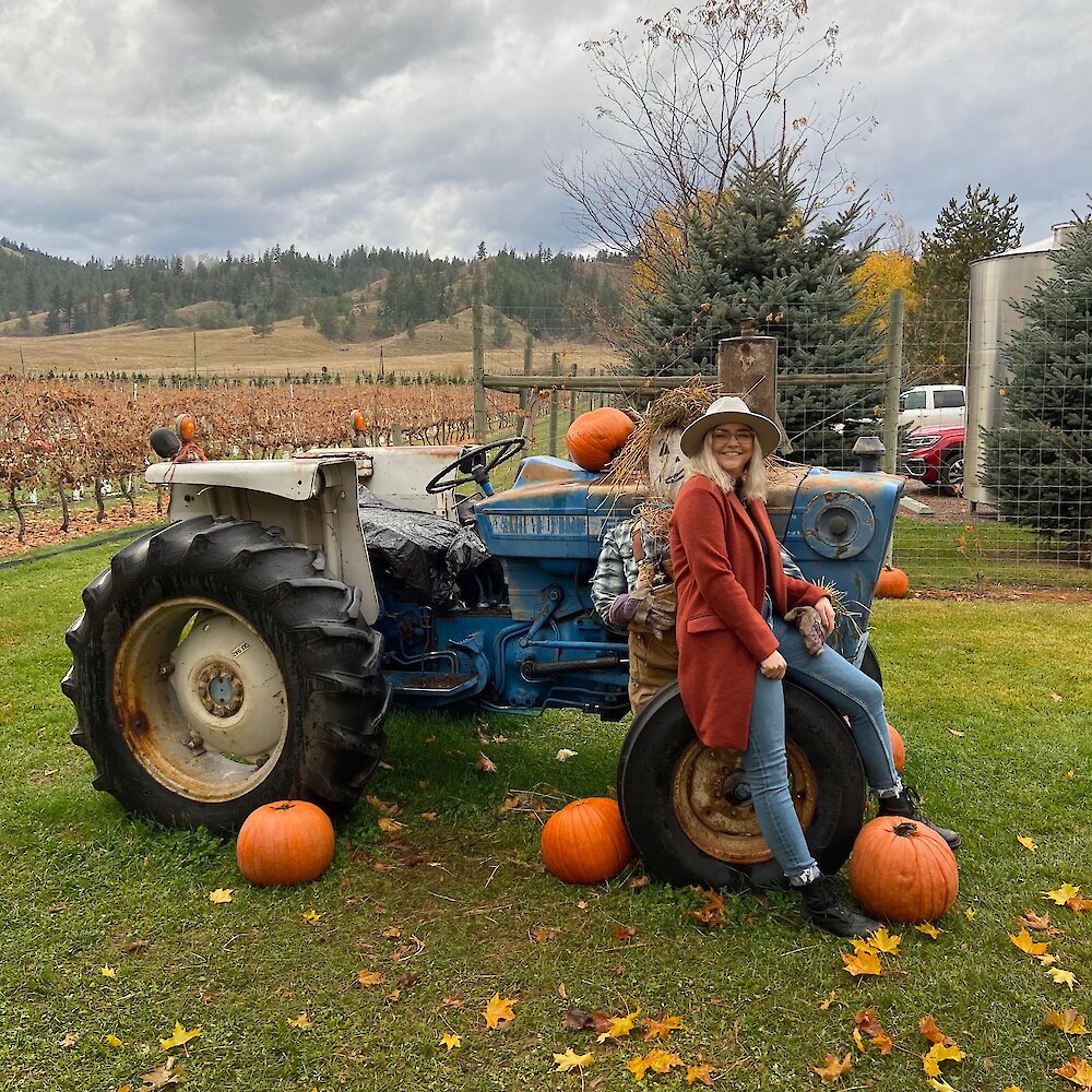 Woman posing on a farm tractor with pumpkins and a fall coloured vines in the background at Privato Vineyard & Winery in Kamloops, British Columbia.