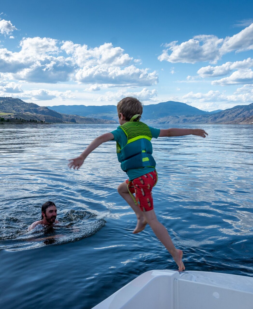 A boy jumping off the boat into Kamloops Lake with sunny blue sky and mountains in the background.