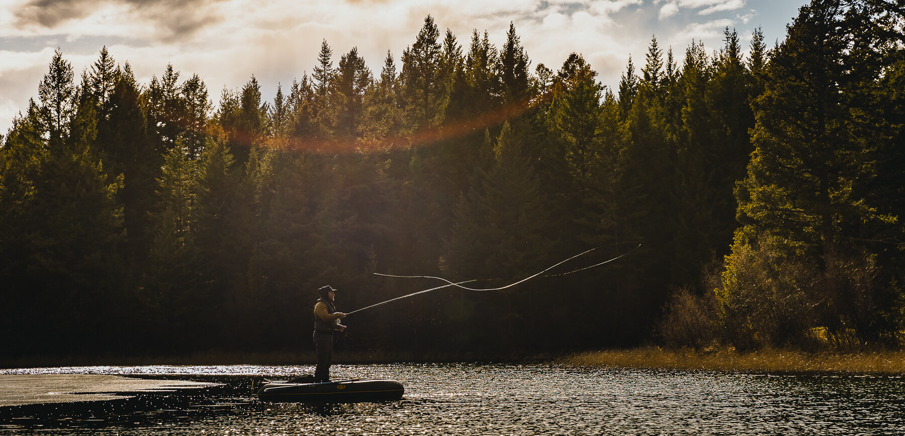 An angler fly fishing from a raft on a sunny lake with trees in the background near Kamloops, BC.