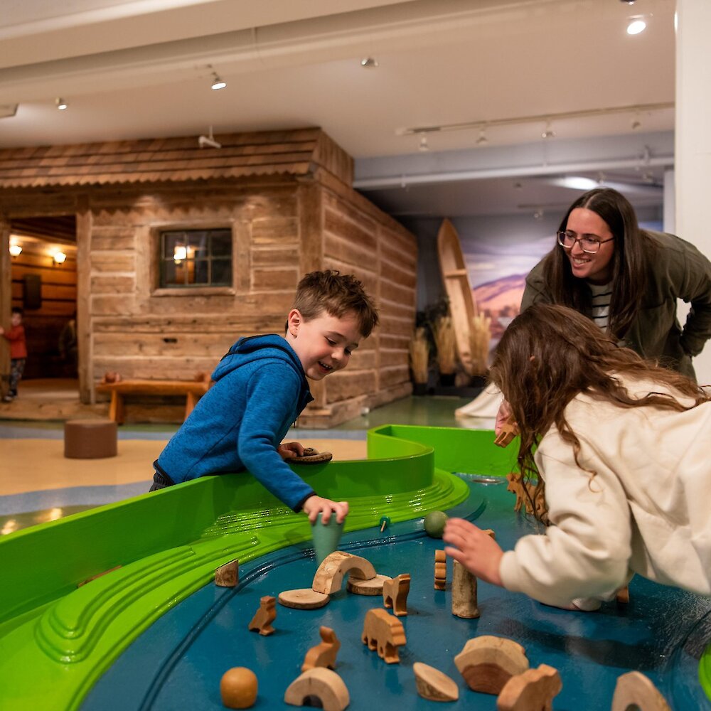 A few children playing with the waterless table with a fur trader's cabin in the background at the Children's Museum in the Kamloops Museum & Archives.