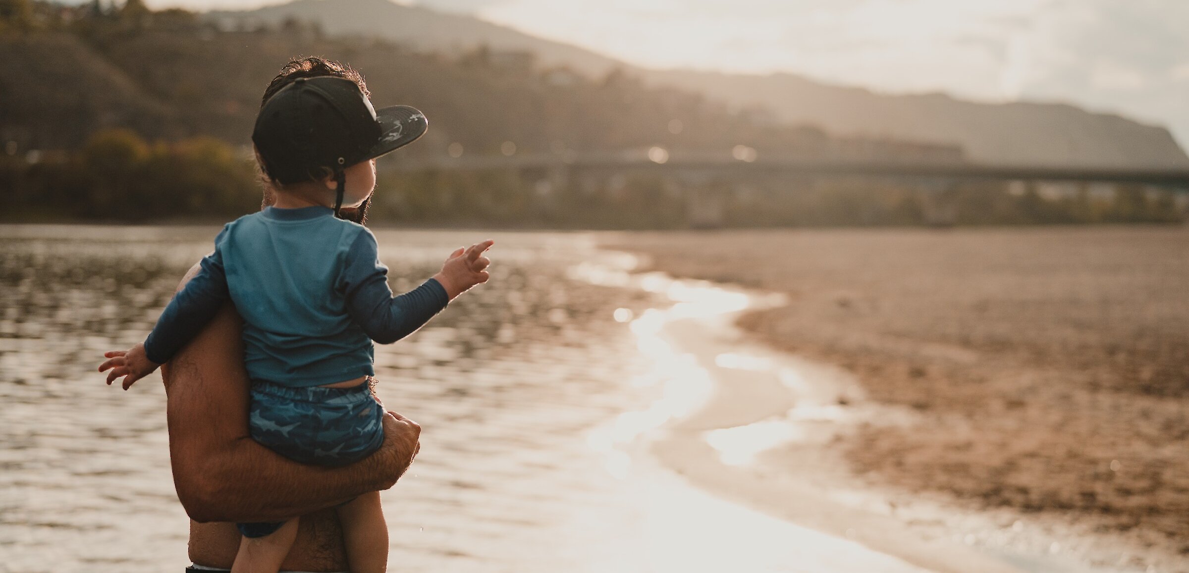 A dad holding his son at Overlander Beach on the North Shore in Kamloops with the mountains and sunny sky in the background.