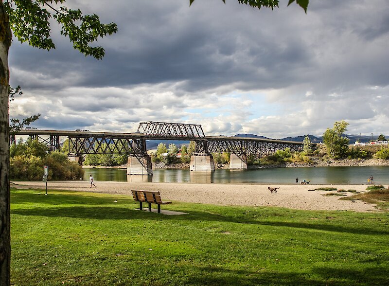 View of Pioneer Park with a beach, benches, and train bridge along the South Thompson River.