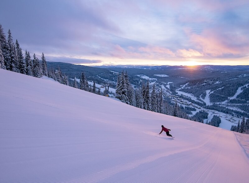 A snowboarder carving down a pristinely groomed trail at Sun Peaks Resort with a view of the mountain's ski trails and sunset in the background.