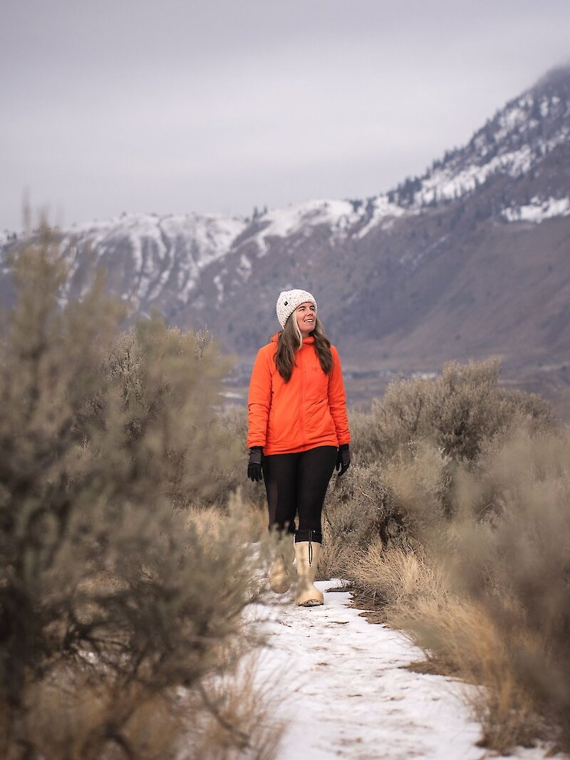 A woman walking through the sagebrush with a snow-covered mountain in the background at Valleyview Nature Park in Kamloops, BC.